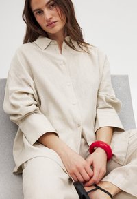 Camicia in lino beige chiaro con bottoni sulla parte anteriore, maniche larghe. Un bracciale rosso su un polso e sandali neri visibili ai piedi.