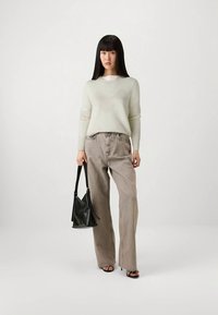 Γυναίκα στέκεται φορώντας ανοιχτό μπεζ πουλόβερ, χαλαρές γκρι τζιν, μαύρες ψηλοτάκουνες σάντες, και κρατώντας μια μαύρη δερμάτινη τσάντα μπροστά σε λευκό φόντο.