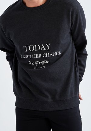 Person iført en sort sweatshirt med hvid tekst på fronten, der lyder "I dag er en ny chance for at blive bedre Etableret 1976".