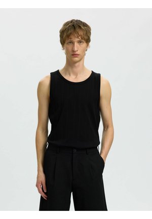 Joven con cabello corto y castaño que lleva una camiseta negra sin mangas con textura de canalé y pantalones negros de cintura alta, de pie con una mano en el bolsillo.