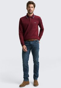Langarm-Poloshirt in Bordeauxrot mit kontrastierendem Kragen, kombiniert mit blauen Jeans und braunen Wildleder-Stiefeletten. Verfügt über ein gesticktes Logo auf der Brust.