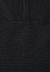 Schwarzer Strickpullover mit hohem Kragen und halbem Reißverschluss. Der Stoff hat ein strukturiertes, geripptes Muster.