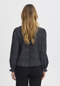 Blusa nera con pois bianchi, maniche a sbuffo e polsini arricciati. Tessuto morbido con una vestibilità comoda, vista da dietro.