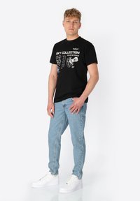 Schwarzes Grafik-T-Shirt mit weißen Texten und Designs, kombiniert mit hellblauen Jeans und weißen Sneakers. Legere Passform und aus Baumwollmaterial.