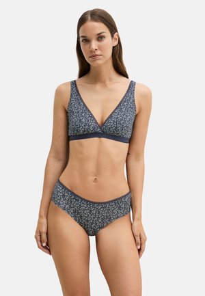 Femme portant un ensemble soutien-gorge et culotte assortis à imprimé floral bleu marine, debout devant un fond blanc uni.