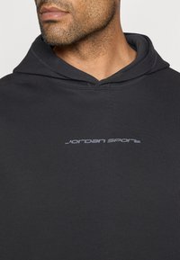 Μαύρη κουκούλα φτιαγμένη από υφασμάτινο υλικό, με καγκουρό τσέπη και διακριτό λογότυπο "jordan sport" σε γκρι χρώμα στο στήθος.