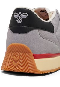 Graue Wildleder-Sneaker mit einer schwarzen Leder-Fersenkappe, die ein Logo trägt, roten Akzenten und einer strukturierten Gummi-Sohle.