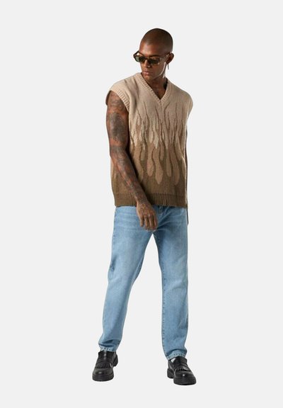Gilet lavorato a maglia beige con motivo a fiamma, scollatura rotonda e design senza maniche, abbinato a jeans azzurri chiari e scarpe nere.