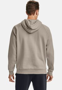 Uomo che indossa una felpa beige e pantaloni neri visto da dietro su uno sfondo chiaro e uniforme.