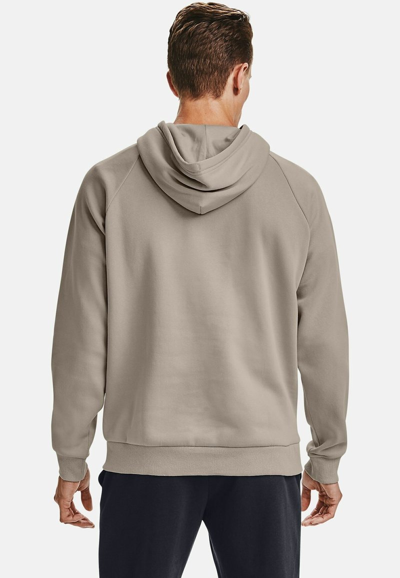 Uomo che indossa una felpa beige e pantaloni neri visto da dietro su uno sfondo chiaro e uniforme.