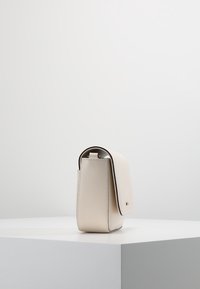 Pieni kermanvärinen nahkalaukku, jossa on sileä pinta, pyöristetty muoto ja läppäkansi. Siinä on minimalistinen muotoilu ja hienovarainen metallinen yksityiskohta.