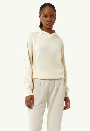 Cremfarbener Rippstrick-Hoodie mit Kordelzug und Raglanärmeln. Kombiniert mit hellbeigen Hosen, die seitliche und frontale Taschen aufweisen.