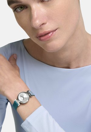 Femeie în bluză albastru deschis care arată un ceas argintiu cu curea bicoloră albastră pe încheietura mâinii stângi, pe fundal alb.