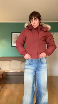 Casaco isolado vermelho com capuz forrado de pelo, fecho à frente e punhos elásticos. Combinado com jeans de perna larga em azul claro com um acabamento desgastado.