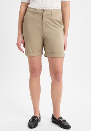 Beige Baumwollshorts mit umgeschlagenen Säumen, ausgestattet mit Vordertaschen und einem Knopfverschluss. Kombiniert mit schwarzen Slippern.