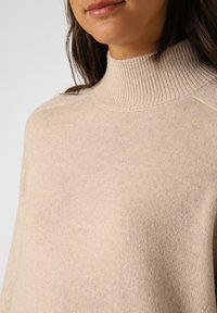 Beige Strickpullover mit hohem Rippkragen, glatter Texture und lässiger Passform, mit dezenten Sprenkeln im Stoff.