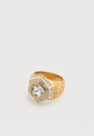 Anello d'oro con design esagonale, caratterizzato da una grande pietra centrale trasparente e da piccole pietre trasparenti circostanti, fascia testurizzata con motivi floreali.