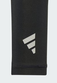 Černé sportovní legíny s texturovaným povrchem, s reflexním stříbrným logem ve tvaru tří diagonálních pruhů blízko lemu.