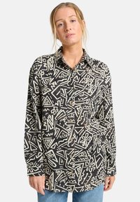 Camisa negra con patrones abstractos blancos, con botones en la parte delantera, mangas largas y cuello. El material parece texturizado, ofreciendo un aspecto casual.