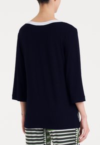 Maglietta blu navy a maniche 3/4 con scollatura a girocollo bianca. Tessuto morbido, vestibilità ampia e design della schiena senza caratteristiche. Indossata sopra pantaloni a righe.