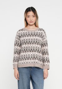 Sweter z dzianiny w miękkim beżu z czarno-białym wzorem w zygzaki, okrągły dekolt i luźny krój, w zestawie z jasnoniebieskimi spodniami z szerokimi nogawkami.