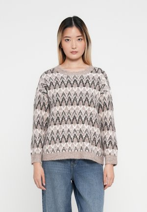 Suéter de punto en suave beige con patrón en zigzag negro y blanco, cuello redondo y corte relajado, combinado con jeans de pierna ancha azul claro.