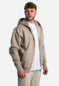 Sudadera beige con cremallera hecha de una tela suave, que cuenta con una capucha amplia, bolsillos delanteros y puños acanalados. La lleva puesta sobre una camisa blanca y pantalones claros.