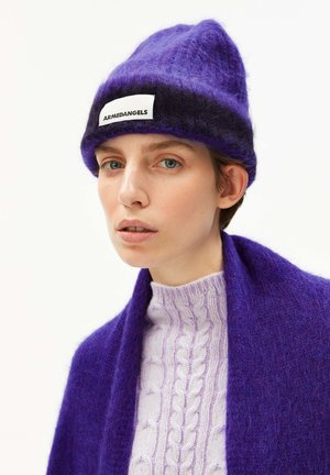 Persoon met een paarse en zwarte fluffy beanie met het label "ARMEDANGELS", een lichtpaarse kabelbreiselsweater en een donkerpaarse fluffy sjaal.