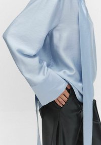 Vaaleansininen silkkipaita, jossa on solmimisdetaili, ilmavat hihat ja leveät hihansuut, yhdistettynä korkeavyötäröisiin mustiin nahkahousuihin. Sujuva tekstuuri.