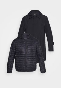 Manteau extérieur noir avec capuche, poches latérales et coupe droite, superposé à une doudoune matelassée noire avec fermeture éclair frontale et capuche.
