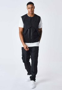 Sort ærmeløs vest med lynlås foran, parret med sorte cargo bukser med flere lommer, båret over en hvid t-shirt og hvide sneakers.
