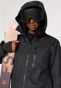 Čierna nepremokavá bunda so zipsom vpredu a nastaviteľnou kapucňou, spárovaná s extra veľkými čiernymi lyžiarskymi okuliarmi a ružovým snowboardom.