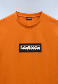 Orange bomulds t-shirt med rund halsudskæring og en sort rektangulær logo-patch med hvid tekst, der står "NAPAPIJRI geographic."