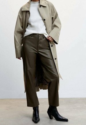 Trench-coat beige porté sur un pull gris, associé à un pantalon en cuir vert olive et des bottines à talons noires. Textures lisses et lignes épurées.