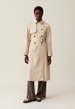 Abrigo beige tipo trench con diseño de doble botonadura, gran cuello y cinturón de lazo. Presenta botones oscuros y una tela suave y texturizada.