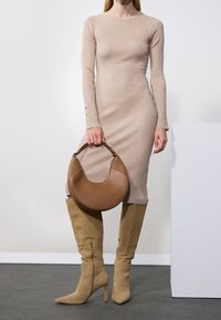 Femme portant une robe côtelée beige et des bottes en daim montant jusqu'aux genoux, tenant un sac à main en cuir marron en forme de croissant.