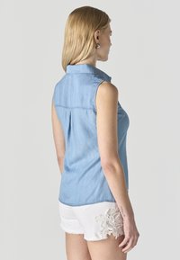Mujer con cabello rubio, vestida con una camisa sin mangas de mezclilla azul clara y pantalones cortos blancos con detalle de encaje, de pie contra un fondo liso.