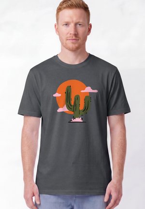 Homme roux portant un t-shirt gris foncé avec un cactus vert et un soleil orange graphique entouré de nuages roses.