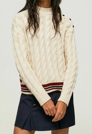 Femme portant un pull crème en maille torsadée avec des détails de boutons et un ourlet rayé, associé à une jupe plissée bleu marine.
