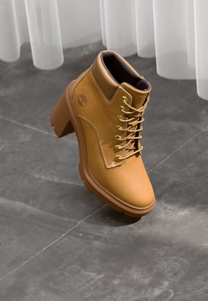 Brune ankelboots med en kraftig hæl, som har gullfargede øyer, hvite sømmer og gummisåle. Glatt lær tekstur, logo på siden.