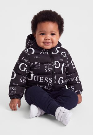 Bébé souriant assis, portant une doudoune noire Guess avec texte blanc, pantalon bleu marine et chaussures blanches sur fond clair.