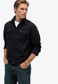 Donkerblauwe button-up shirt met een puntige kraag, twee borstzakken en omgeslagen mouwen, gecombineerd met middelwashes denim jeans en een horloge.