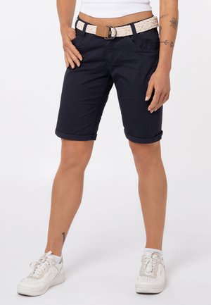 Vrouw draagt marineblauwe knielange shorts met opgerolde zoom, een gevlochten witte riem en witte sneakers, staand met één hand in de zak.