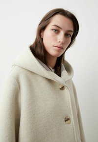 Cappotto di lana beige con un ampio cappuccio, dotato di due grandi bottoni. Texture morbida e design minimalista. Sfondo semplice.
