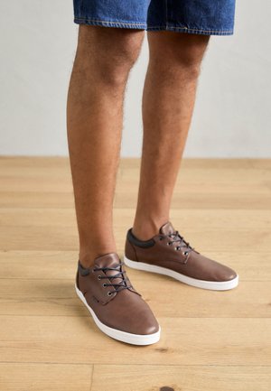 Adidași din piele maro cu vârf rotund, accente negre la guler și șireturi albastre, așezați pe un podea din lemn deschis.