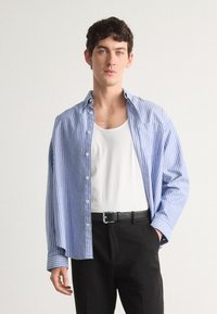 Chemise boutonnée rayée bleu et blanche, coupe décontractée, en coton, à manches longues, associée à un débardeur blanc et un pantalon noir.