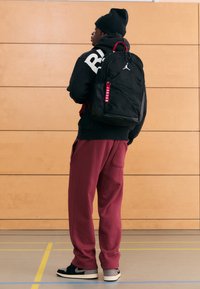 Fekete hátizsák vörös részletekkel és logóval, fekete kapucnis pulóver és bordó melegítőnadrág felett viselve. A háttérben fa fal és tornatermi padló látható.