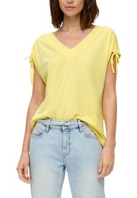 Gelbes V-Ausschnitt-T-Shirt mit kurzen Ärmeln und Bindebändern. Weicher, leichter Stoff kombiniert mit hellblauen hochgeschnittenen Jeans.