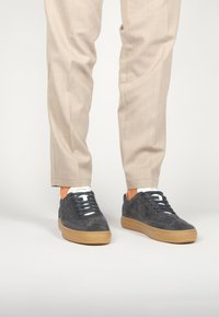 Zapatillas de ante gris con suela de goma beige, cordones blancos y un logotipo sutil, combinadas con pantalones beige claros de corte ajustado.