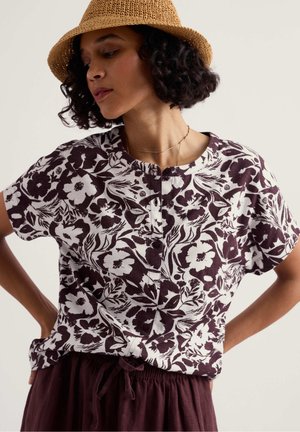 Žena s hnedým tkaným klobúkom, krátkymi kučeravými vlasmi, bielou blúzkou na gombíky s tmavým kvetinovým vzorom a tmavohnedými nohavicami so šnúrkou.